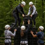 Young people taking part in a crate stack challenge activity in woodland, working in a team to build and balance on a rising tower of crates while wearing safety harnesses.